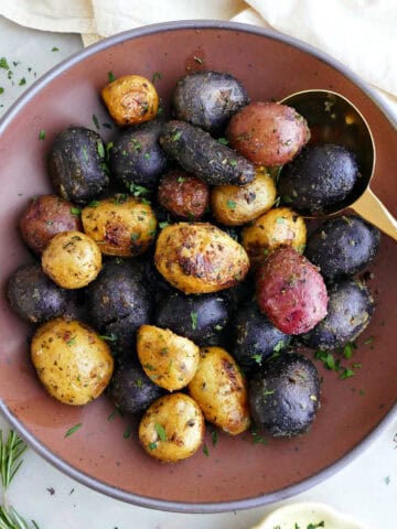 Roasted potatoes in a serving bowl with a spoon next to herbs and a napkin.