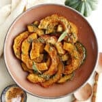 Roasted acorn squash in a serving bowl next to spices, spoons, squash stem, and napkin.