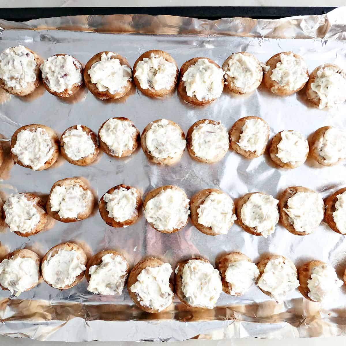Cremini mushroom caps stuffed with boursin cheese on a foil lined baking sheet.