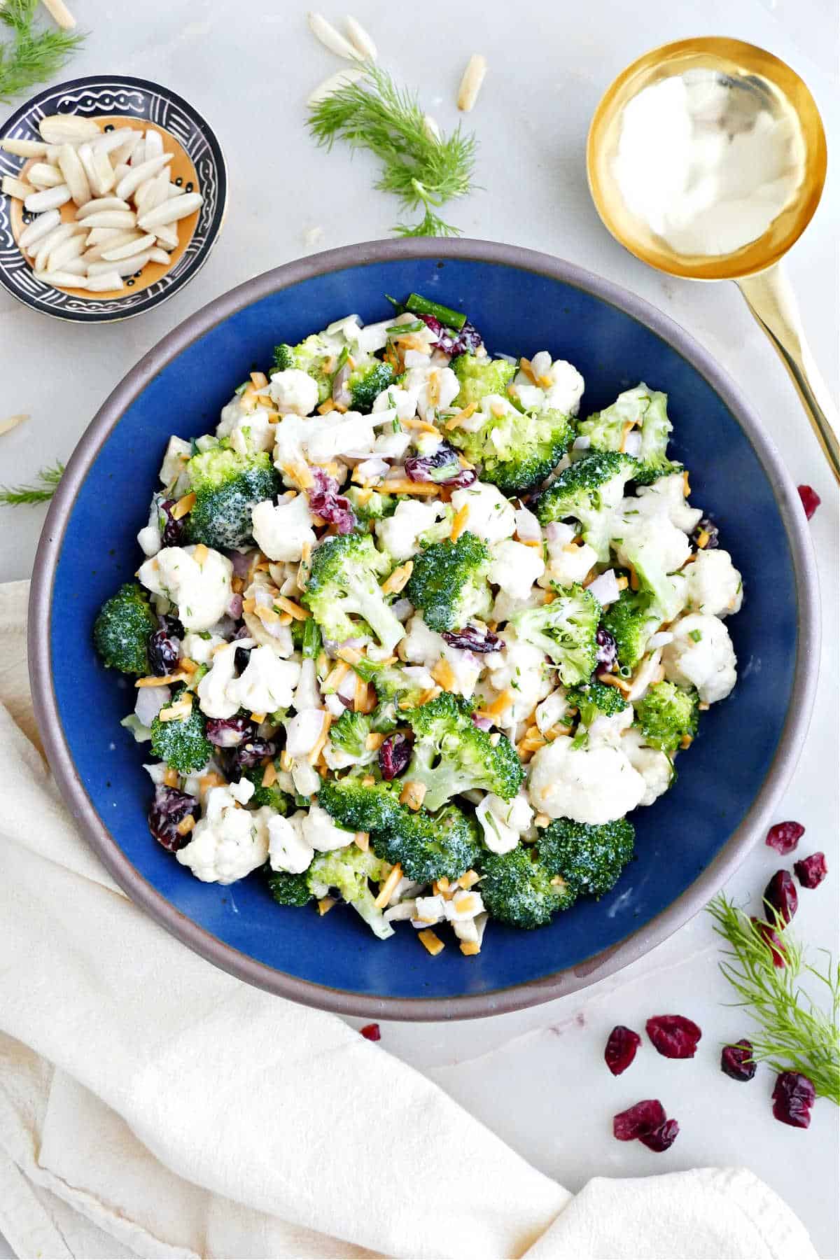 Broccoli and cauliflower salad in a bowl next to garnishes, a napkin, and a spoon.