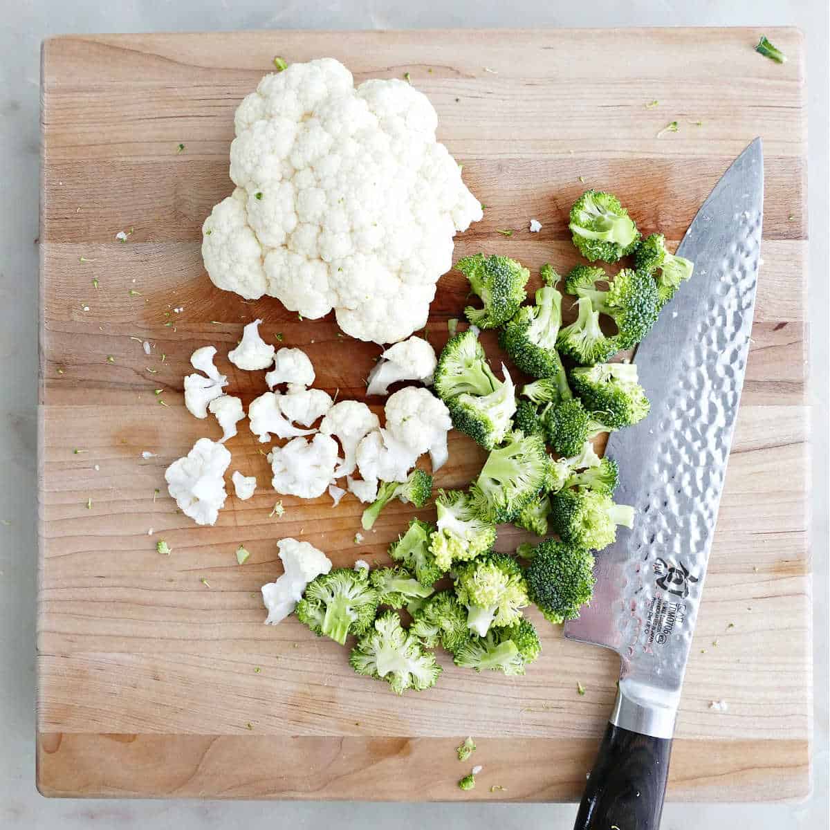 Broccoli and cauliflower being sliced into florets on a cutting board with a knife.