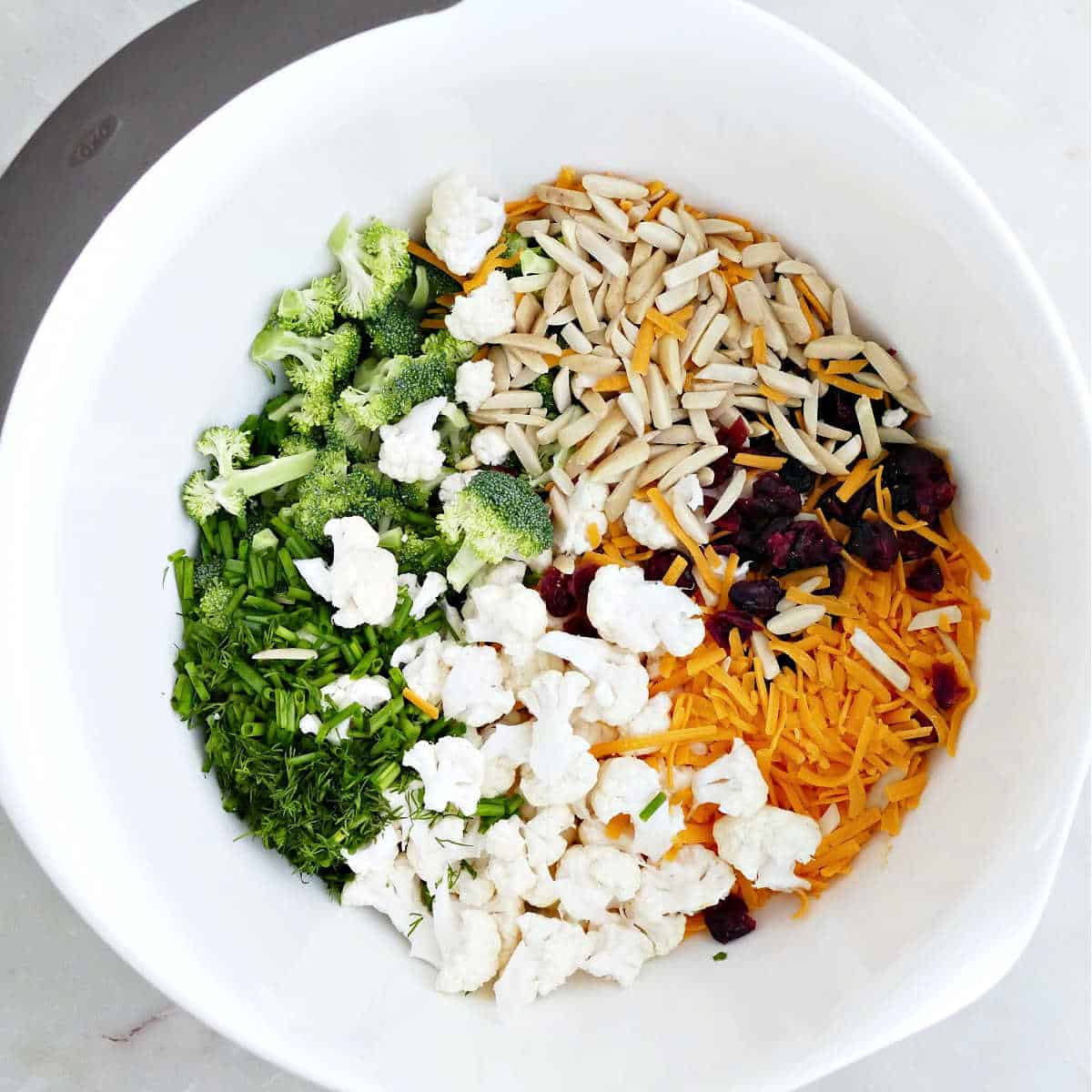 Herbs, broccoli, cauliflower, almonds, dried cranberries, and cheddar cheese in a bowl.