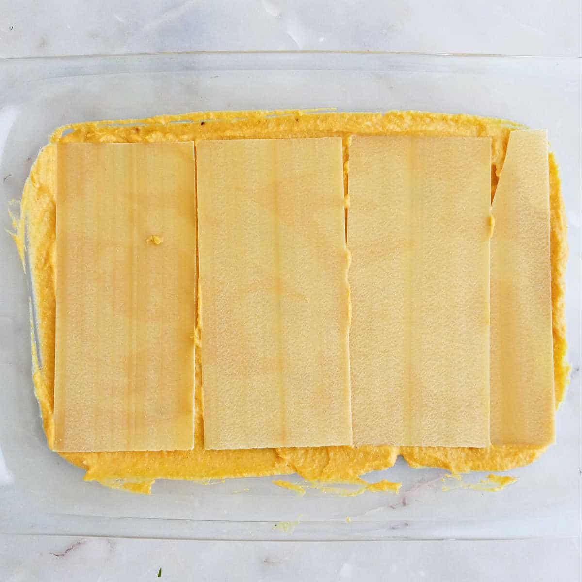 A baking dish with butternut squash sauce on the bottom topped with lasagna noodles.
