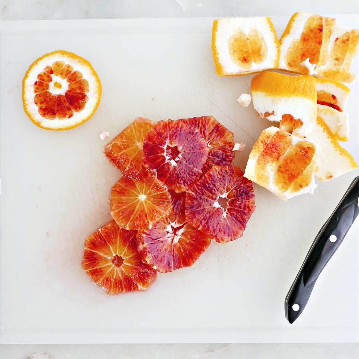 Blood oranges cut into thin slices on a cutting board next to a knife.