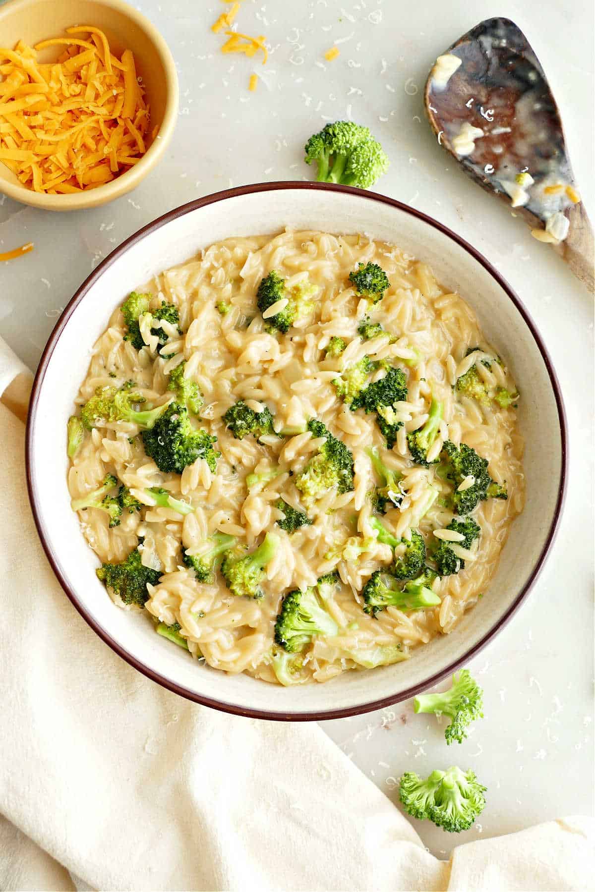 Broccoli orzo in a serving bowl next to napkin, spoon, and bowl of cheese.