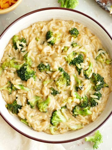 Broccoli orzo in a serving bowl next to napkin, spoon, and bowl of cheese.