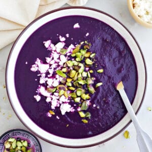 Purple sweet potato soup topped with pistachios and feta in a bowl with a spoon.