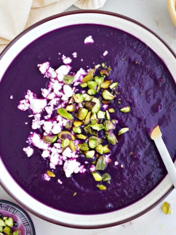 Purple sweet potato soup topped with pistachios and feta in a bowl with a spoon.