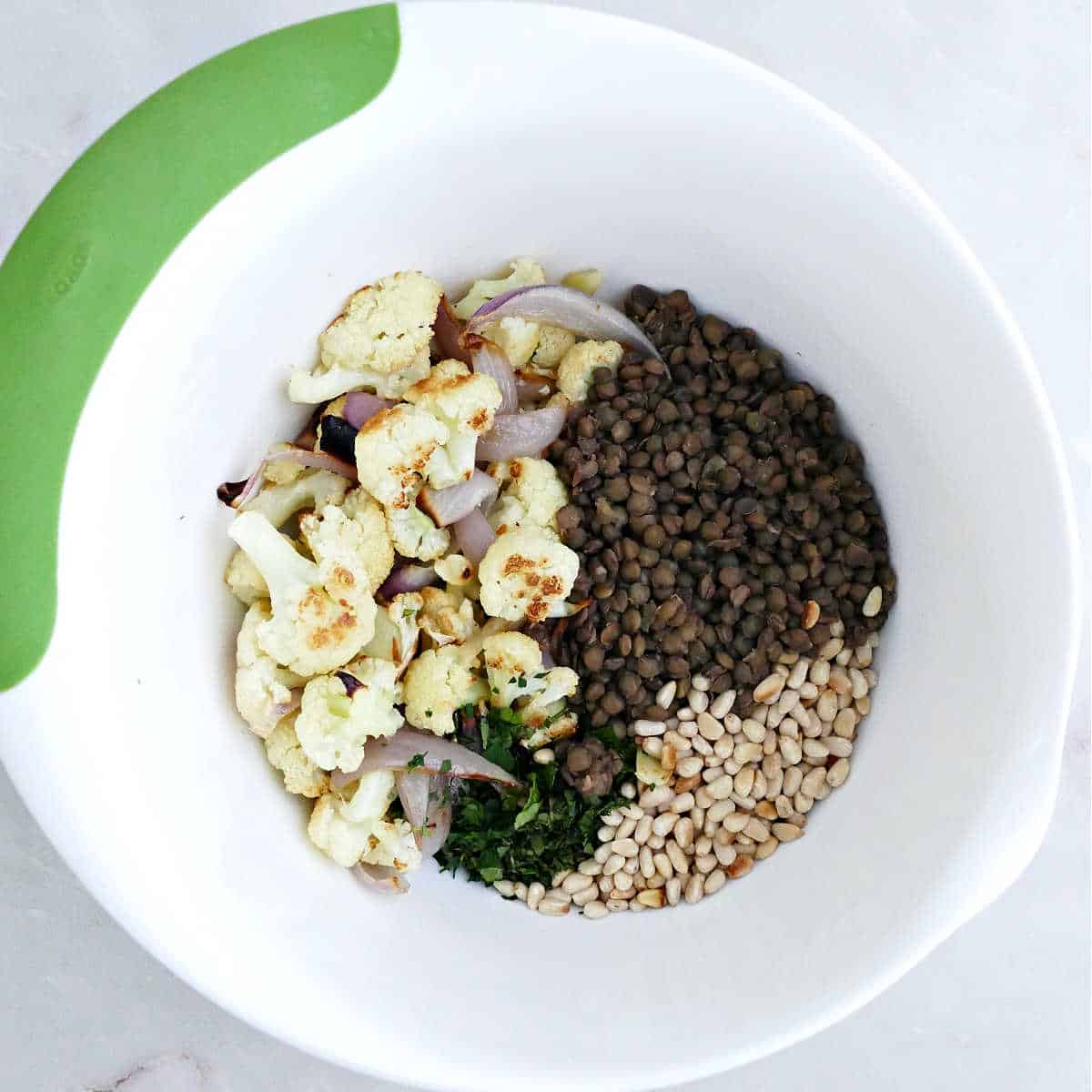 Roasted cauliflower and onion, lentils, pine nuts, and herbs in a mixing bowl.