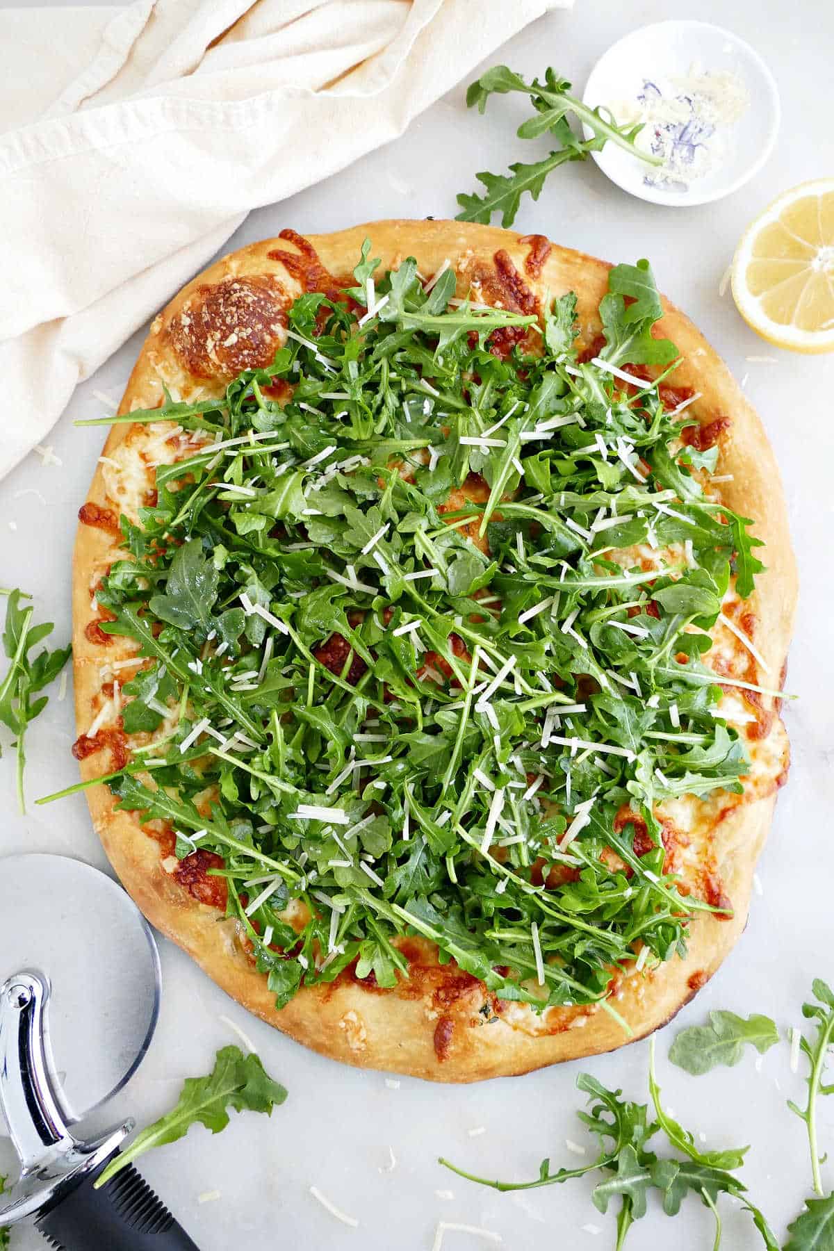 Arugula pizza on a counter next to ingredients, a pizza cutter, and a napkin.