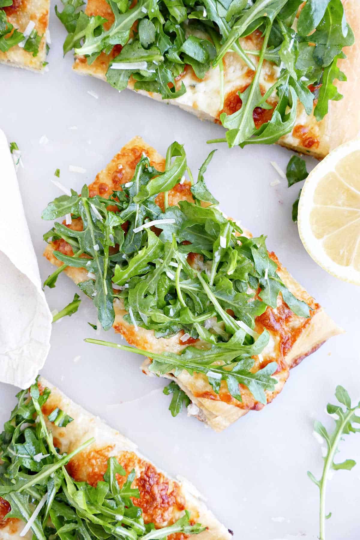 Slice of white pizza topped with arugula next to other pieces and a lemon.