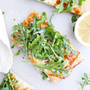 Slice of white pizza topped with arugula pizza next to other pieces and a lemon.