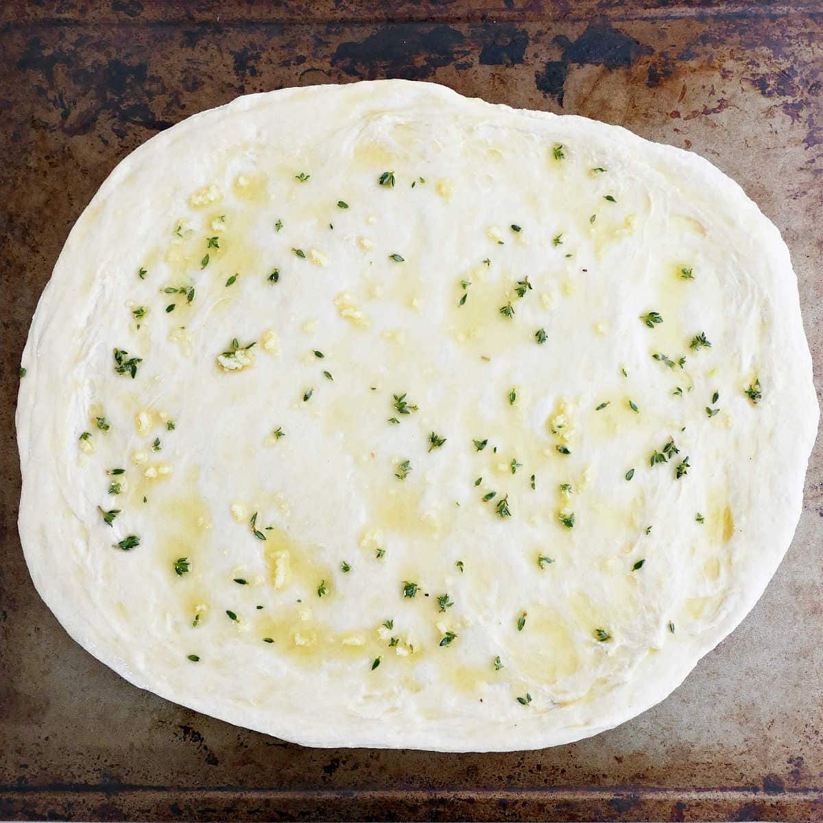 Pizza dough brushed with olive oil, garlic, and thyme on a baking sheet.