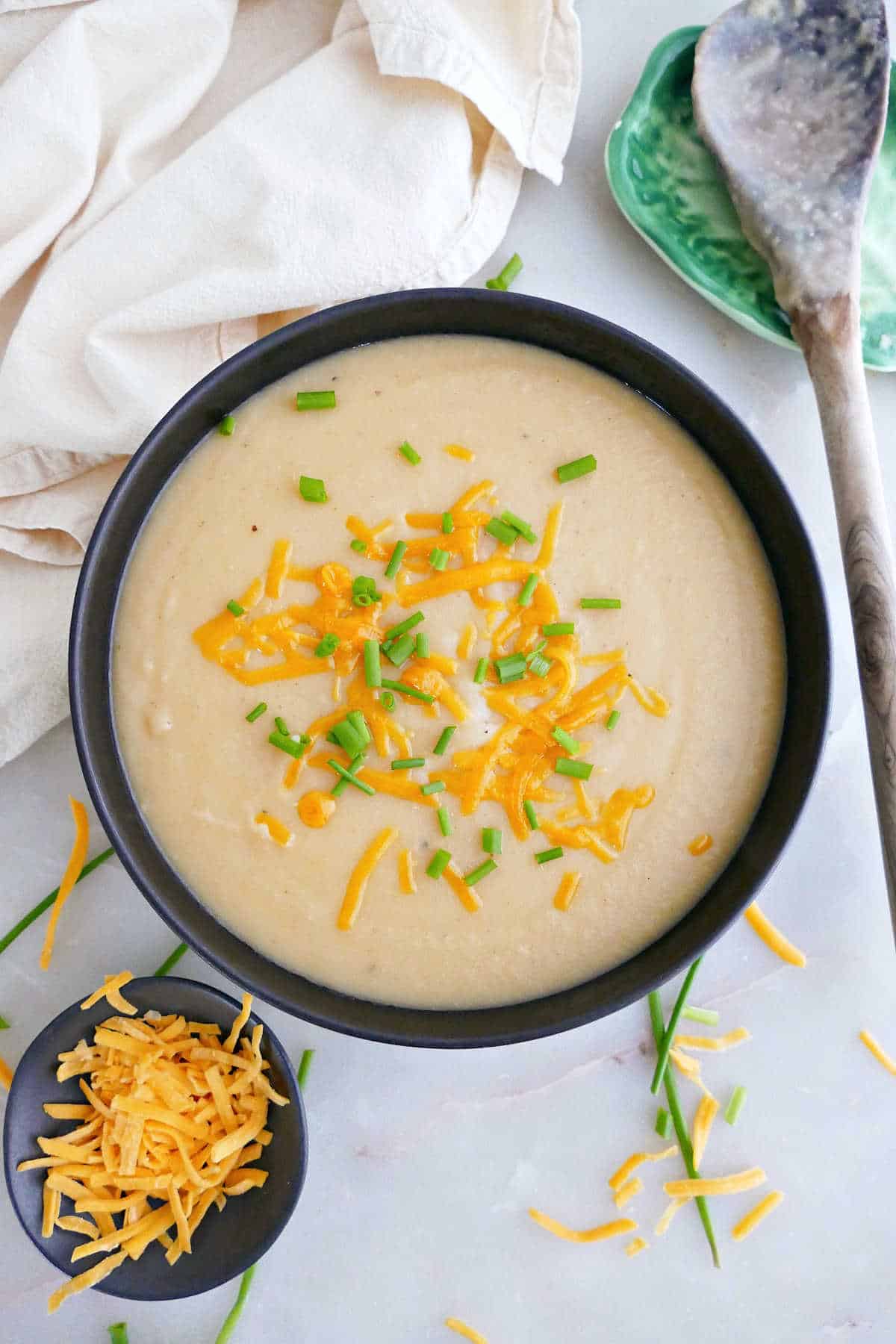 Potato soup with chives and cheese on a counter next to toppings, spoon, and napkin.