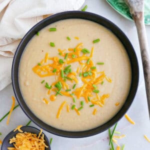 Potato soup with chives and cheese on a counter next to toppings, spoon, and napkin.