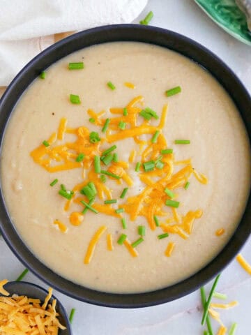 Potato soup with chives and cheese on a counter next to toppings, spoon, and napkin.