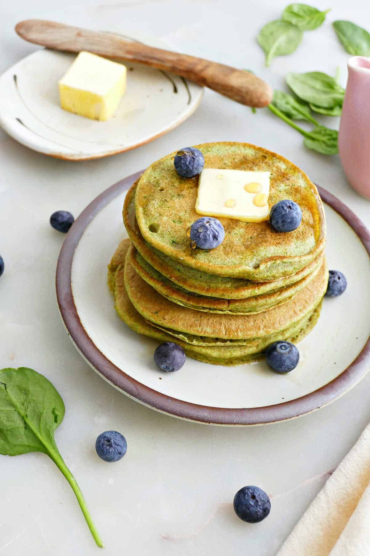 Stack of spinach banana pancakes with butter and blueberries on a plate.