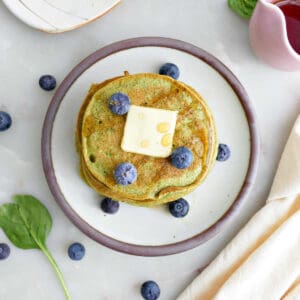 Spinach banana pancakes with butter, syrup, and blueberries on a plate surrounded by ingredients and napkin.