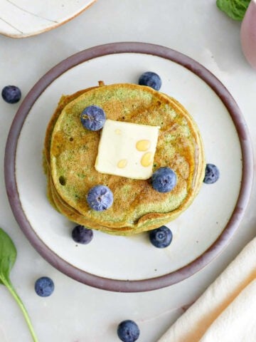 Spinach banana pancakes with butter, syrup, and blueberries on a plate surrounded by ingredients and napkin.