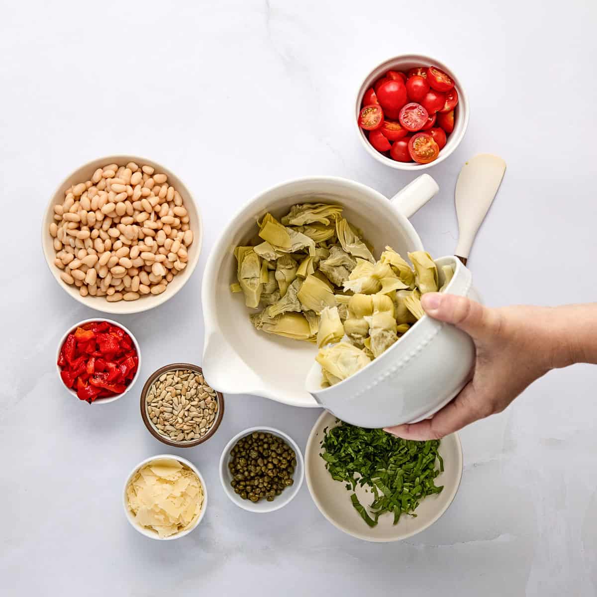 Woman's hand pouring artichoke hearts into a bowl surrounded by other ingredients.