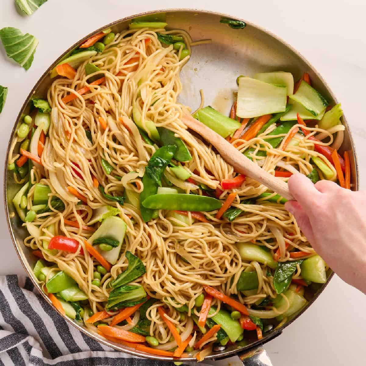 Woman stirring noodles and vegetables in a skillet to coat them in sauce.