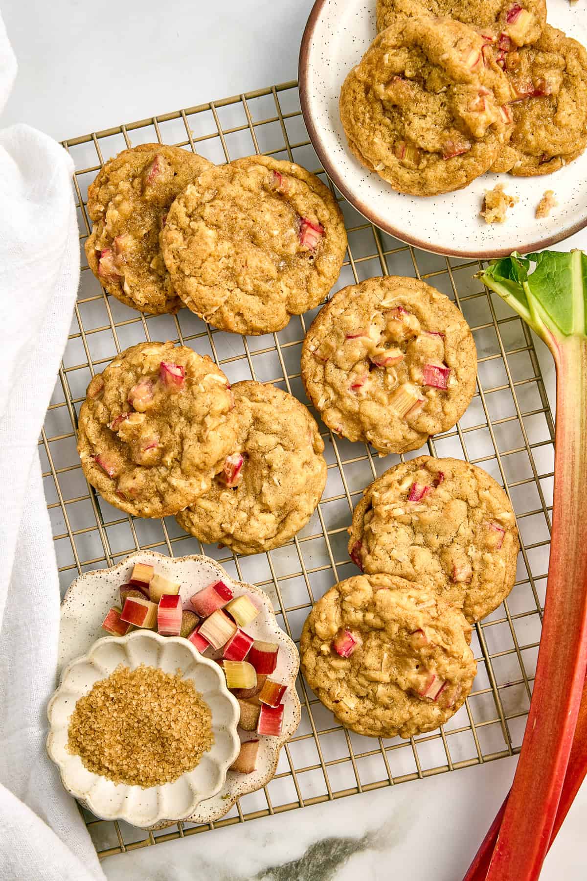 Seven rhubarb cookies on a wire rack next to ingredients, plate of cookies, and rhubarb stalk.