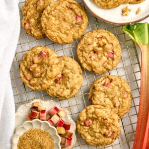 Seven rhubarb cookies on a wire rack next to ingredients, plate of cookies, and rhubarb stalk.
