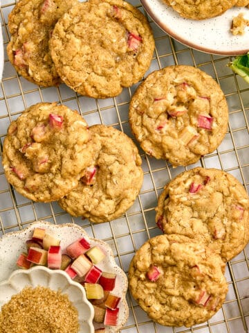 Seven rhubarb cookies on a wire rack next to ingredients, plate of cookies, and rhubarb stalk.