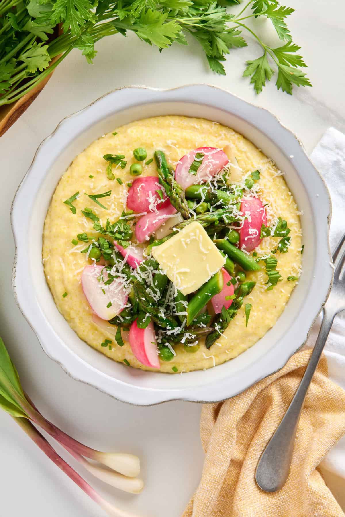 Bowl of polenta topped with spring vegetables, butter, and cheese next to ingredients, napkin, and fork.