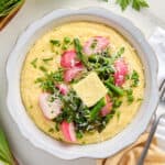 Bowl of polenta topped with spring vegetables, butter, and cheese next to ingredients, napkin, and fork.