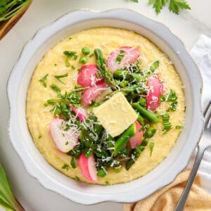 Bowl of polenta topped with spring vegetables, butter, and cheese next to ingredients, napkin, and fork.