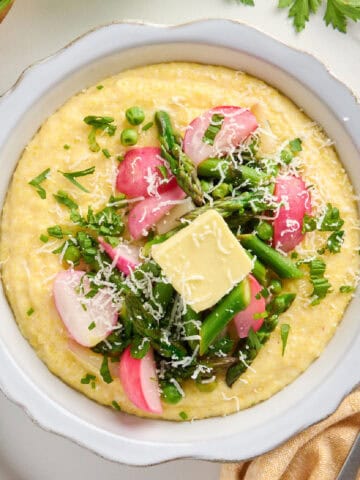 Bowl of polenta topped with spring vegetables, butter, and cheese next to ingredients, napkin, and fork.