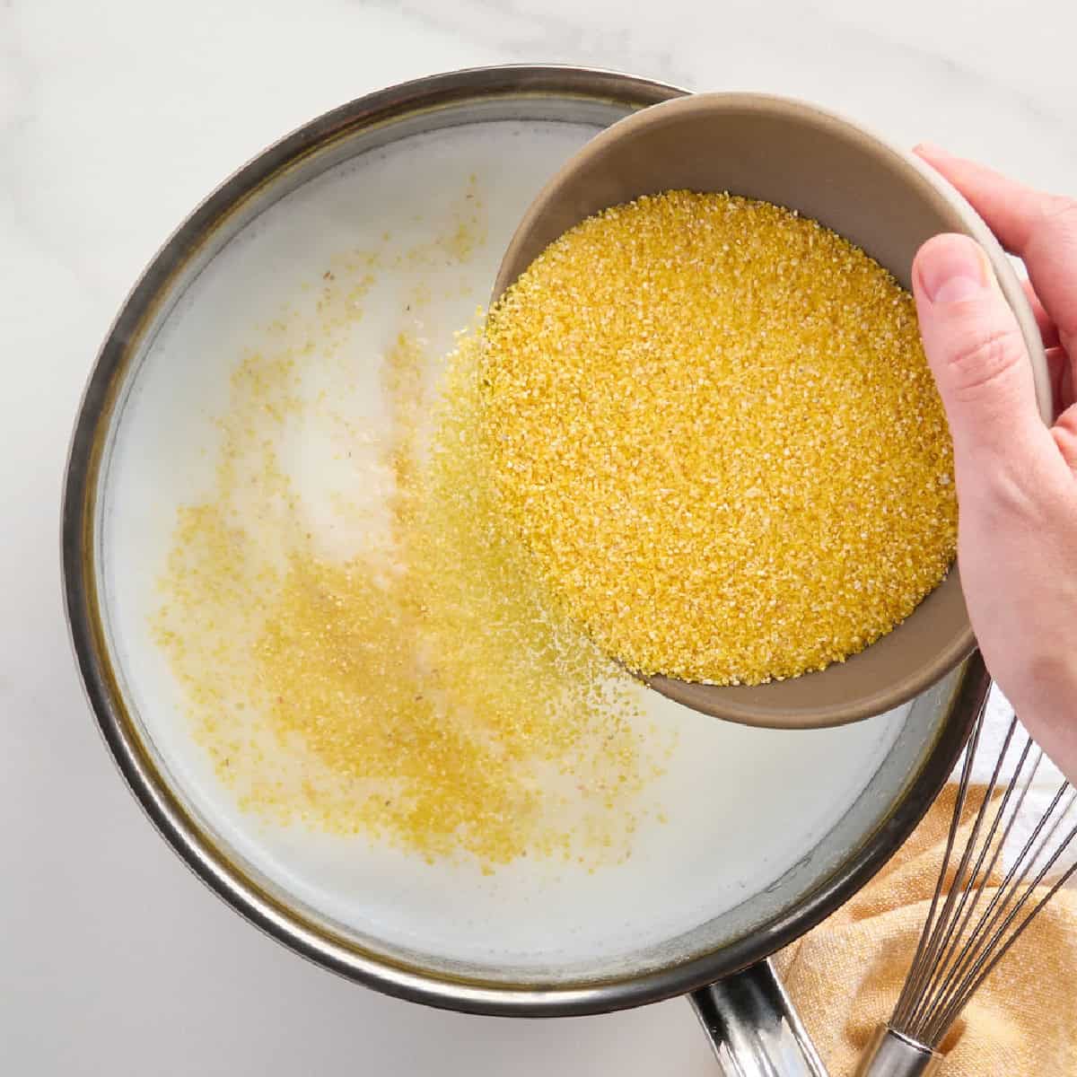 Dry polenta being poured into a pot with milk and water on a stove.