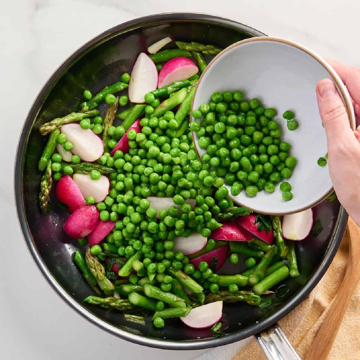 A woman adding peas to a skillet with other spring vegetables.