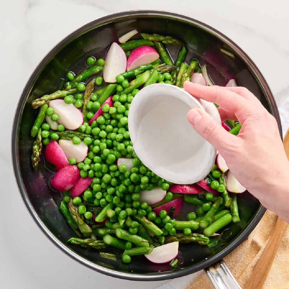 A woman adding water to spring vegetables cooking in a skillet.