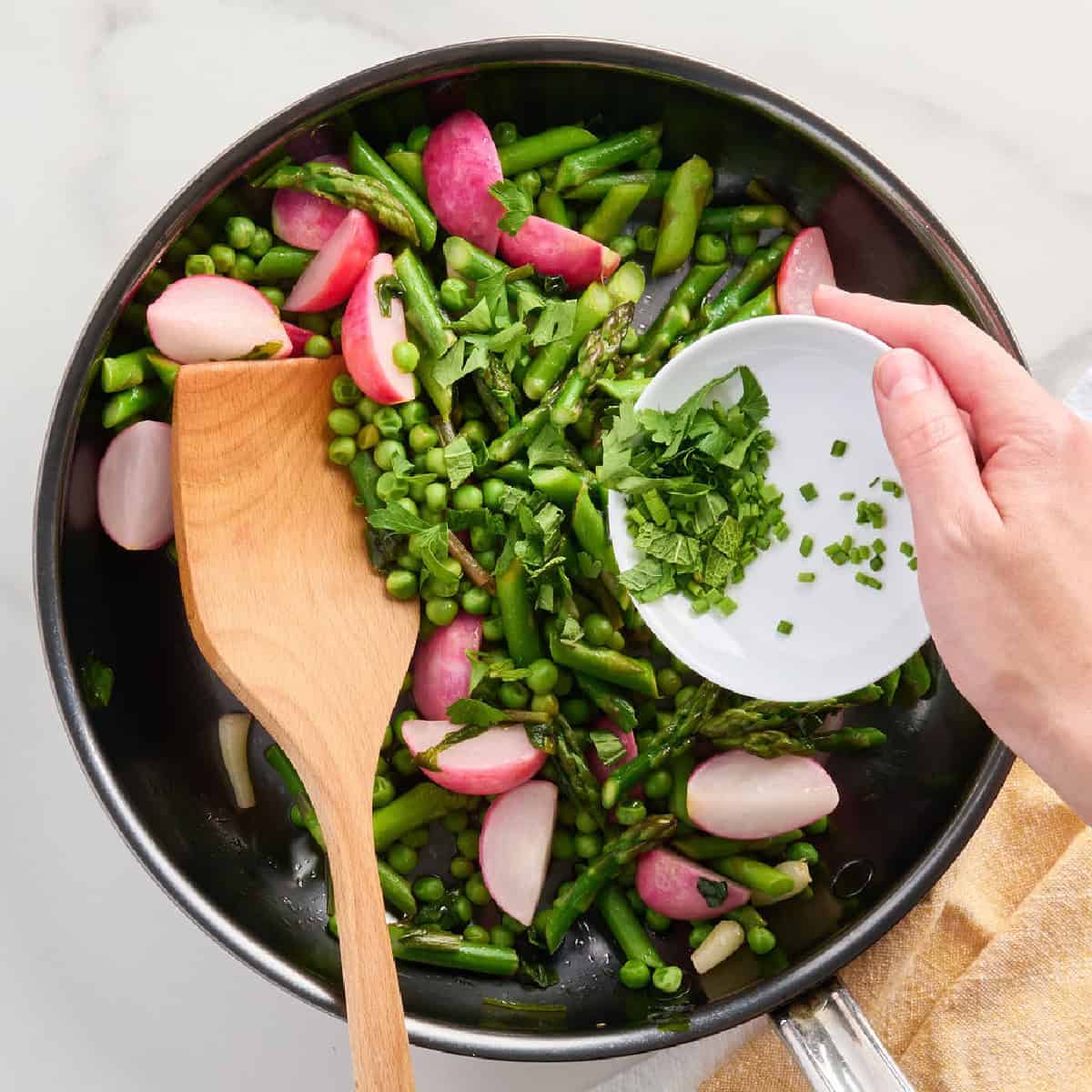 A woman adding fresh herbs to a skillet of cooked vegetables.