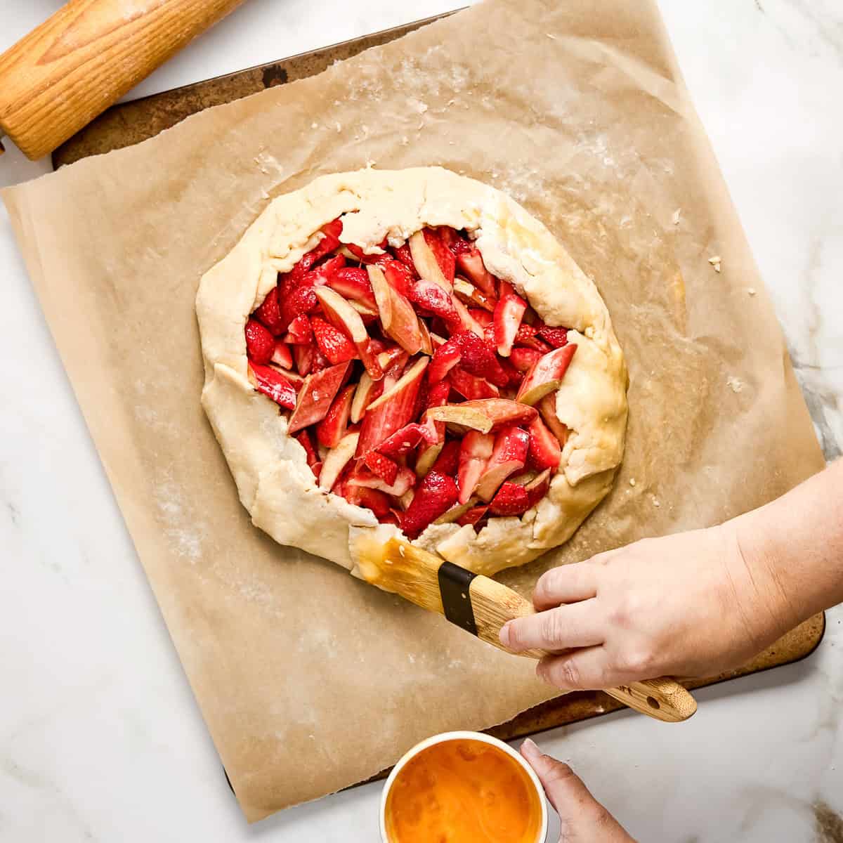 Woman brushing the crust of a strawberry rhubarb galette with a beaten egg.