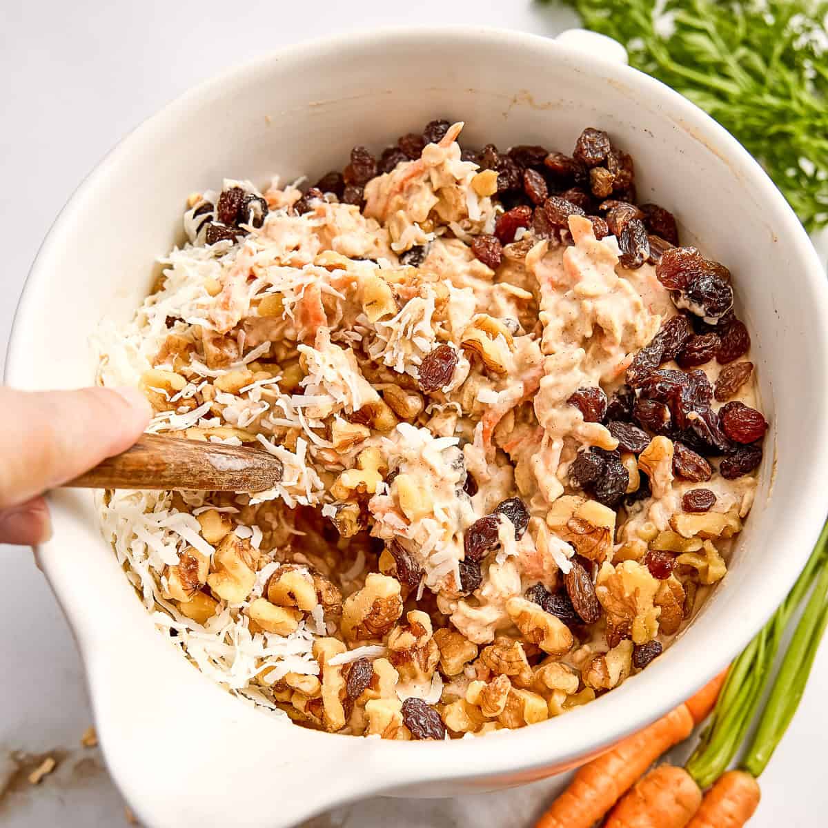 Woman folding coconut, raisins, and walnuts into oatmeal batter in a bowl with a spatula.