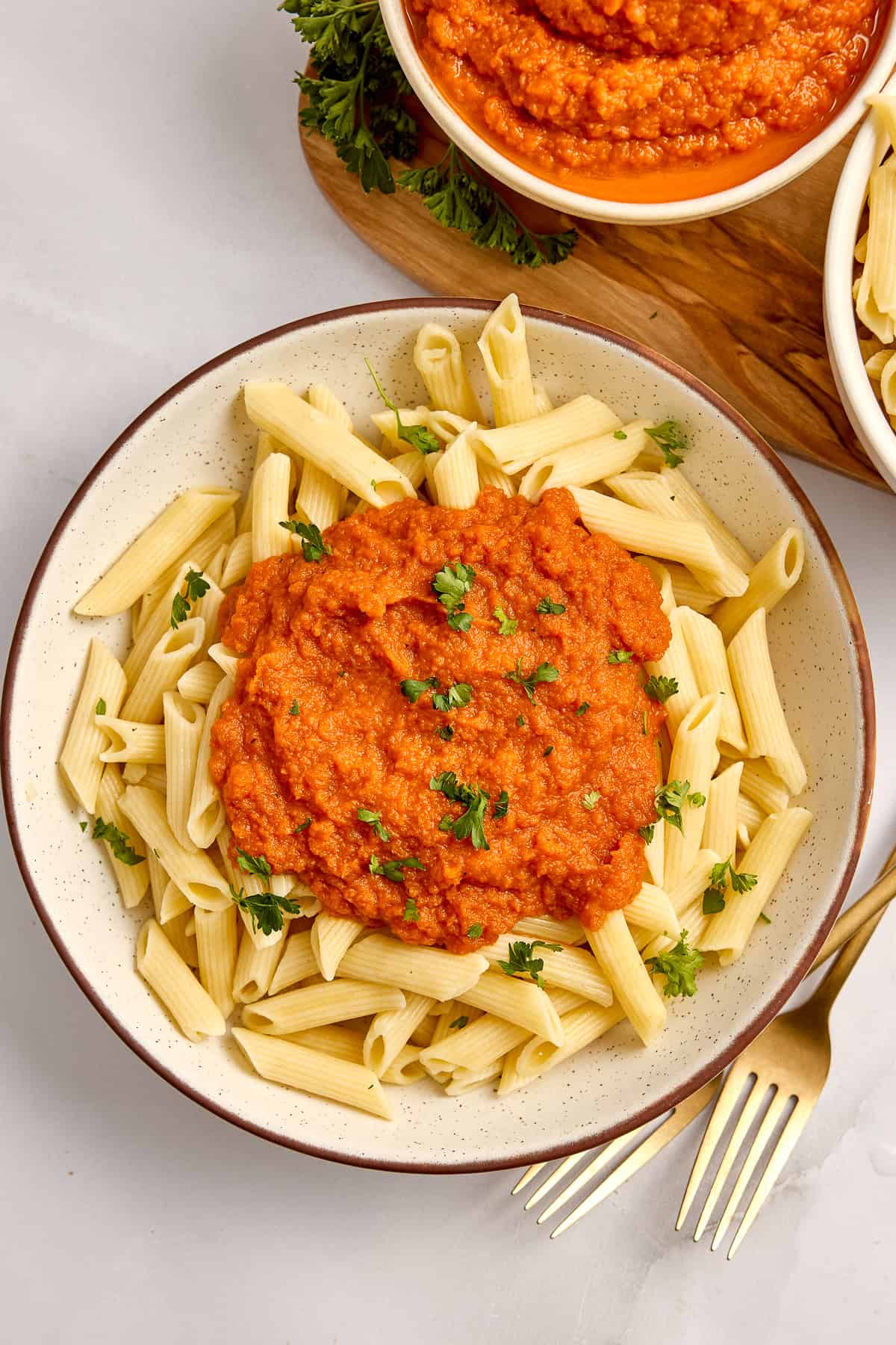 Bowl of penne pasta topped with carrot pasta sauce and fresh parsley next to forks.