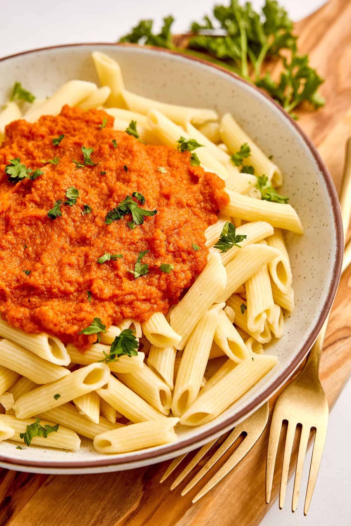 Penne with carrot pasta sauce and fresh parsley in a bowl next to parsley and forks.
