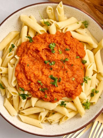 Bowl of penne pasta topped with carrot pasta sauce and fresh parsley next to forks.