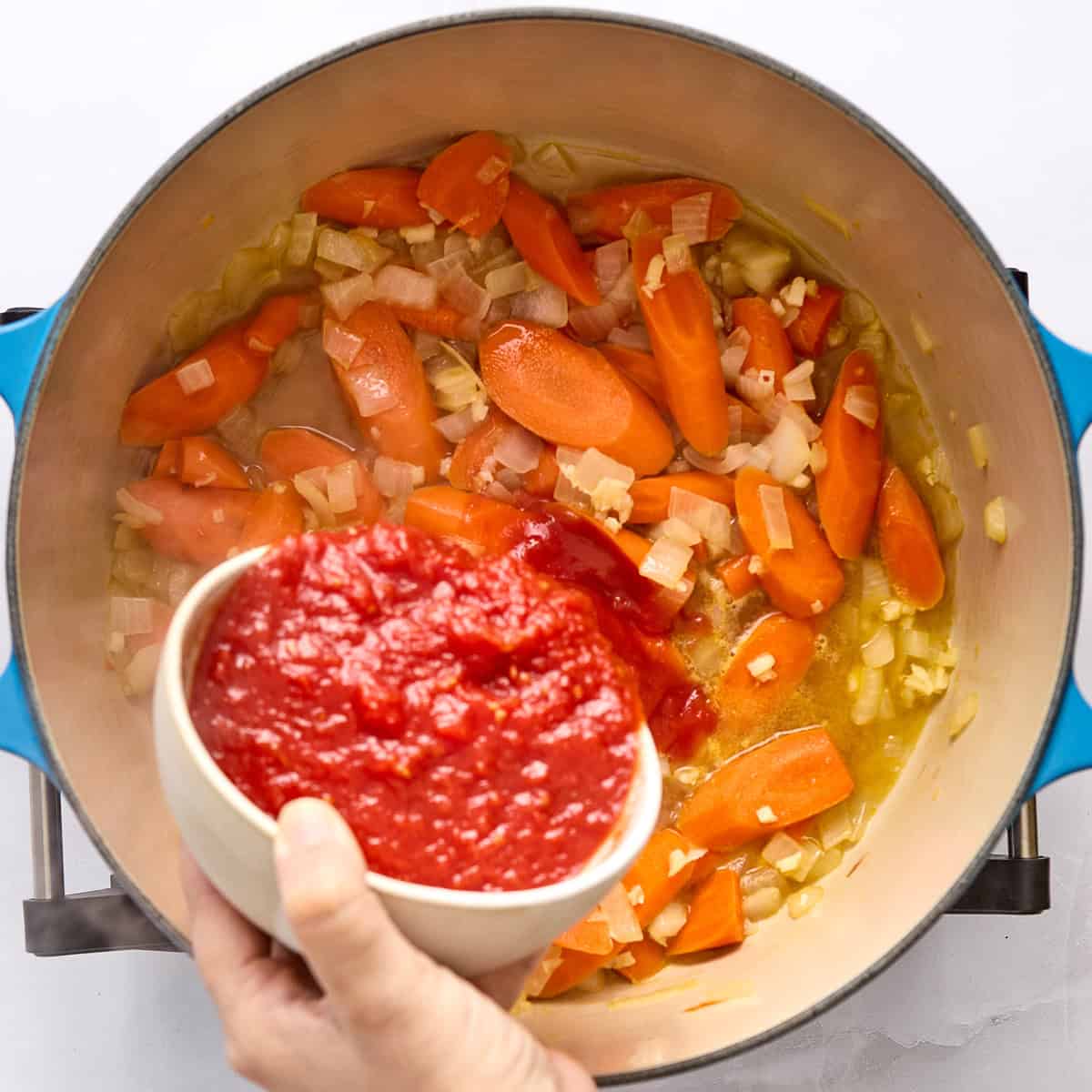 Woman adding crushed tomatoes to a pot with carrots, onions, and garlic cooking in oil.