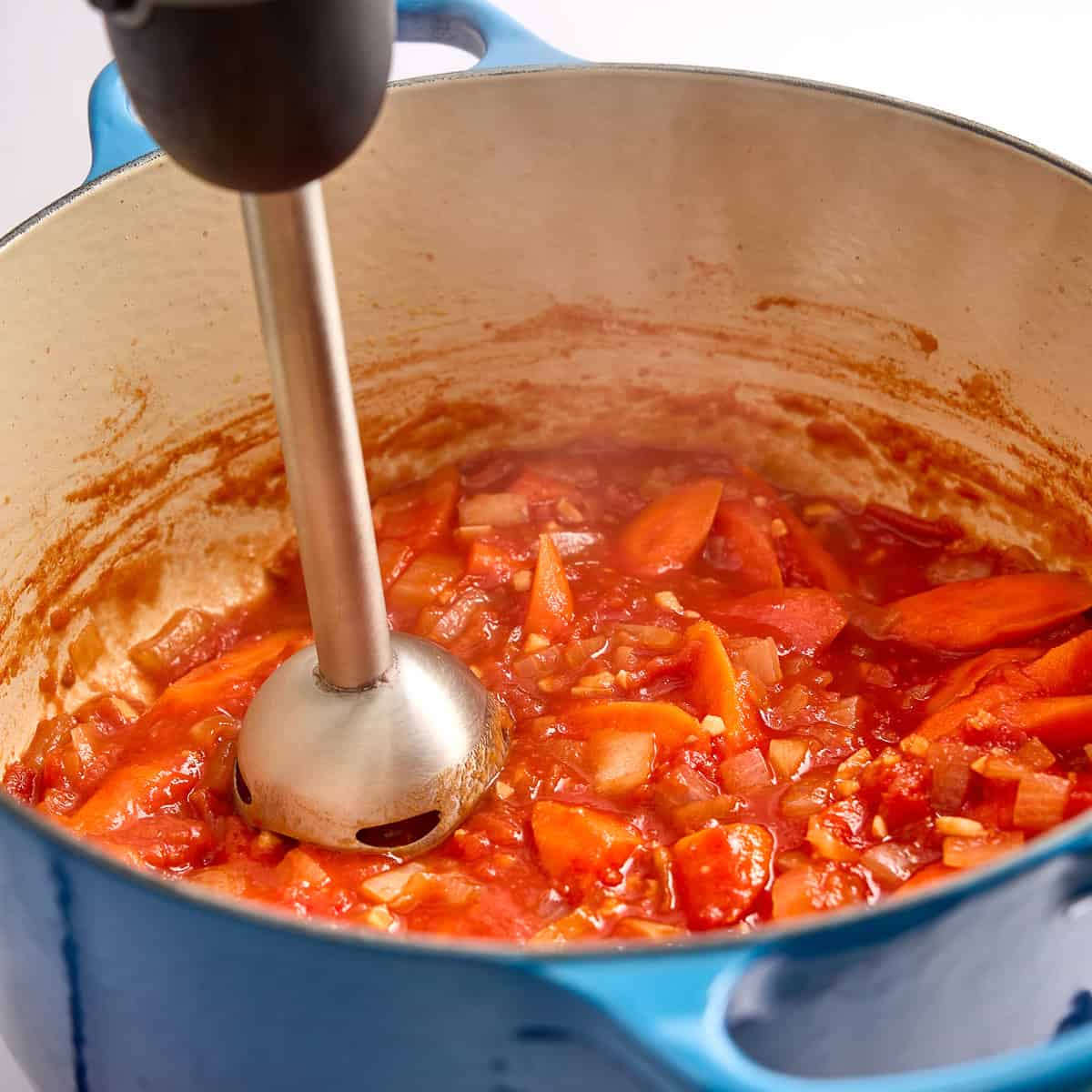 Woman using an immersion blender to blend carrot pasta sauce in a large pot.