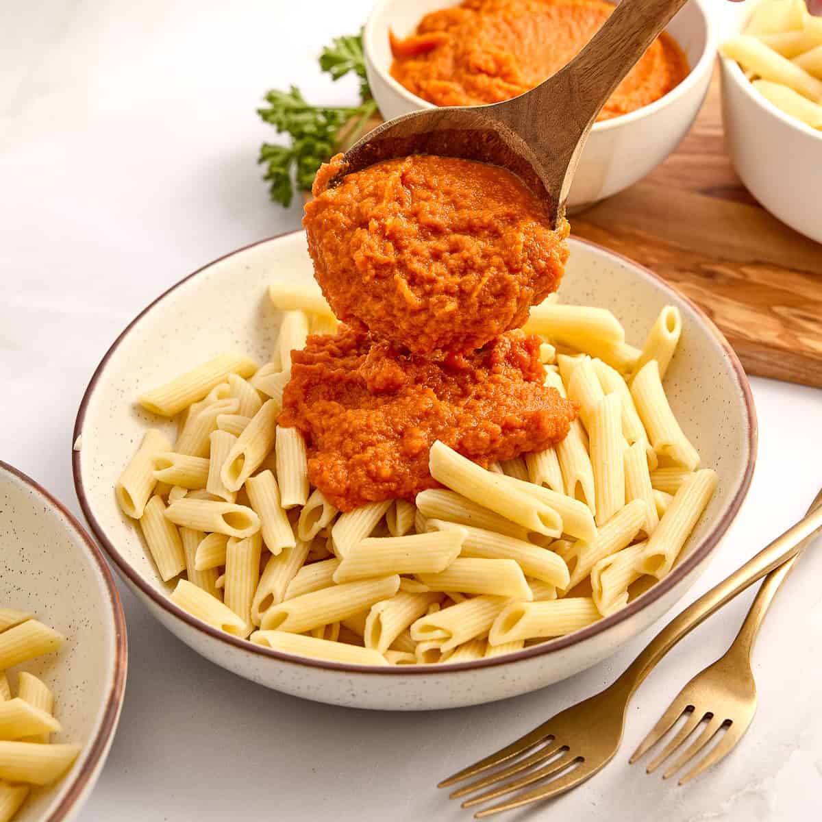 Woman ladling carrot sauce over cooked penne pasta in a bowl next to forks.