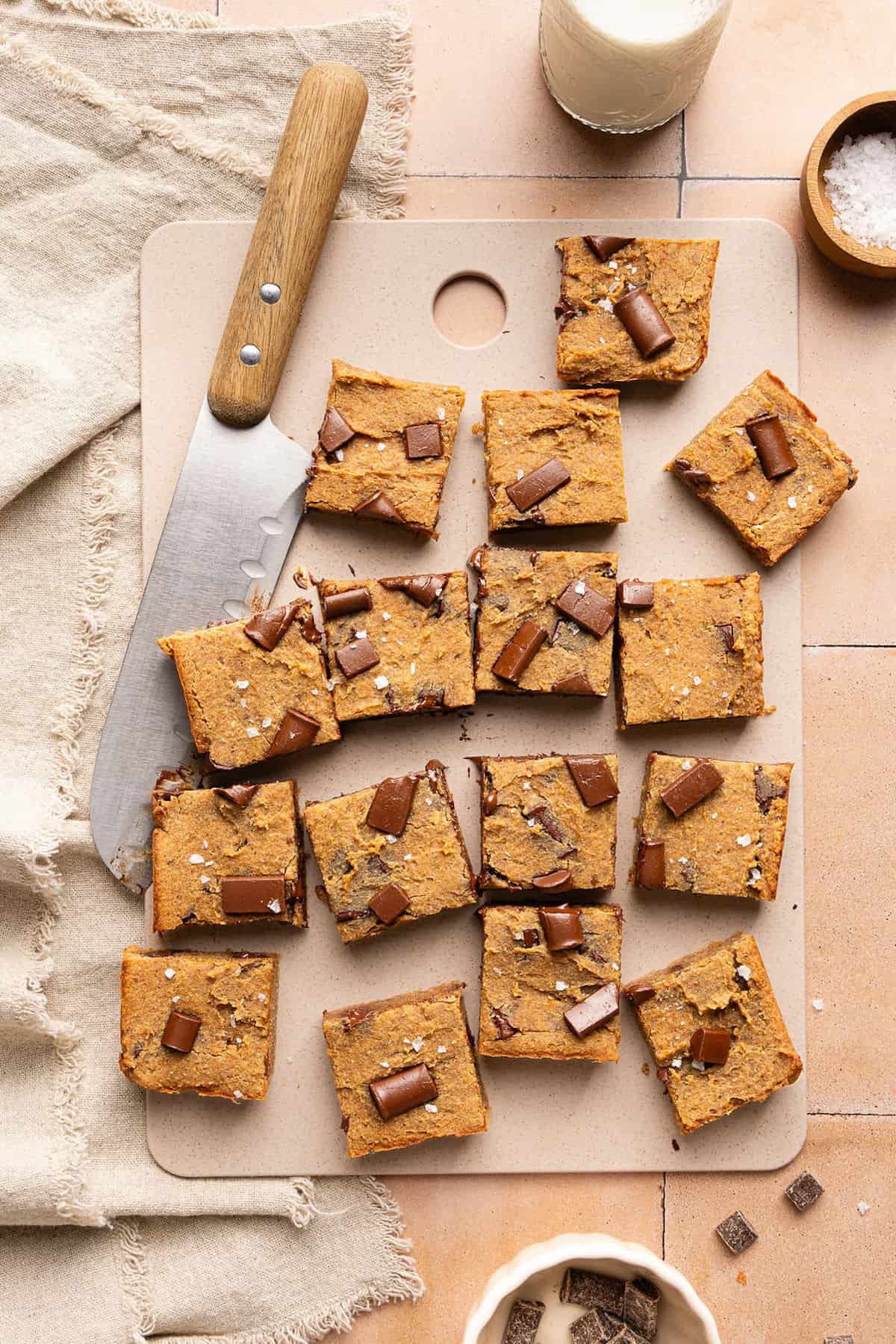 Sixteen blondies with chocolate chunks on a cutting board next to a knife and ingredients.