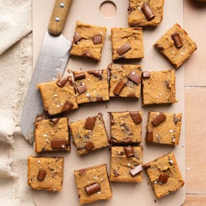 Sixteen blondies with chocolate chunks on a cutting board next to a knife and ingredients.