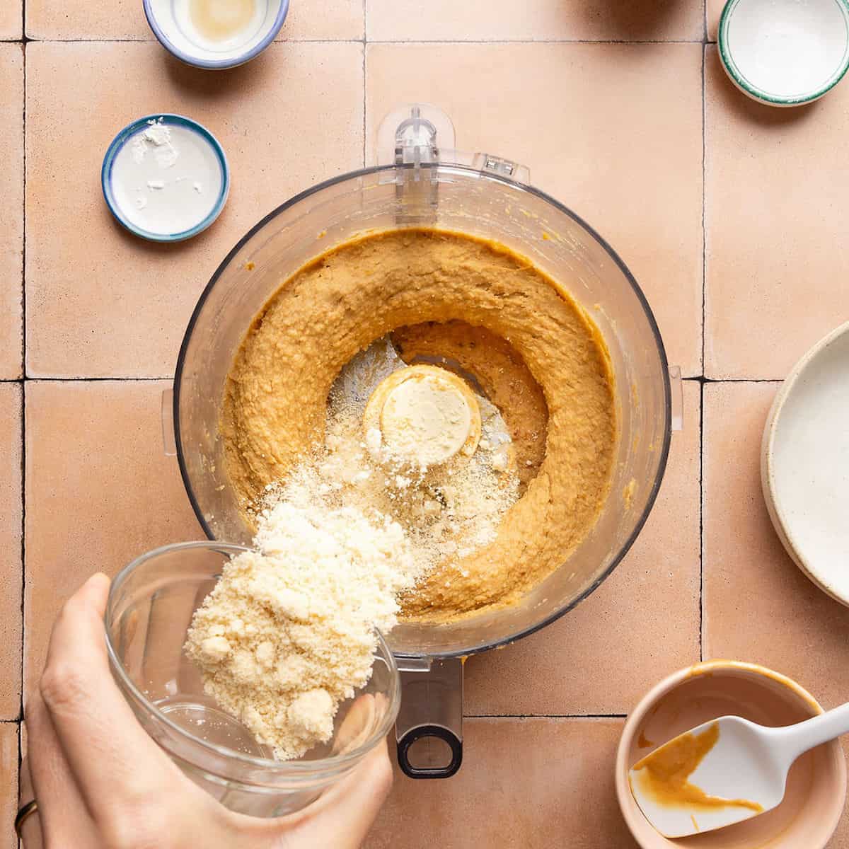 Woman pouring almond flour into a food processor with blended blondie batter.
