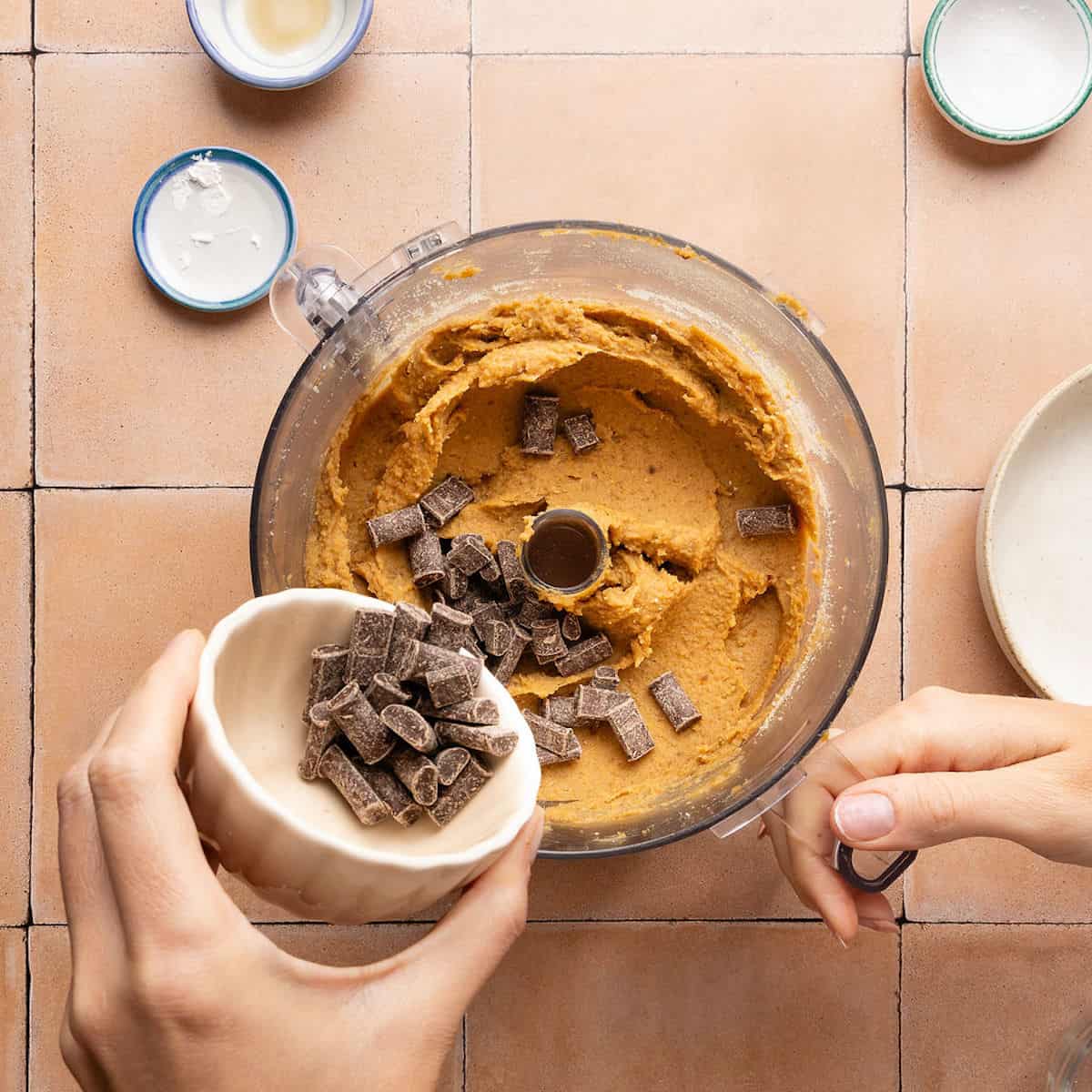 Woman pouring dairy-free chocolate chunks into a food processor with batter for blondies.