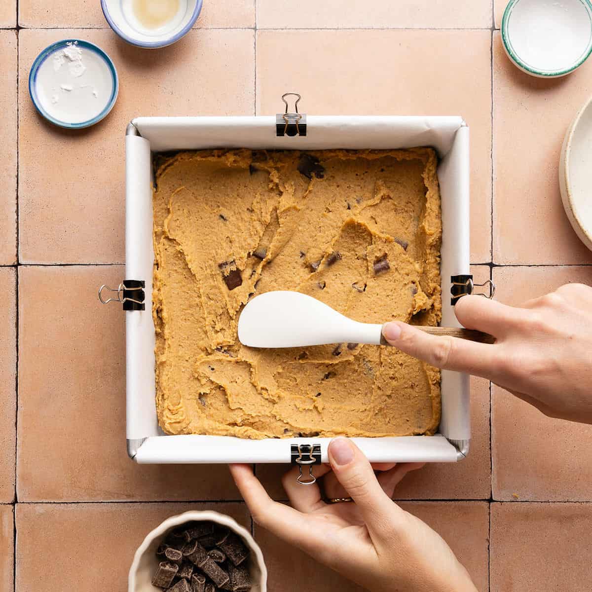 Woman spreading blondie batter with a spatula into a lined square baking pan.