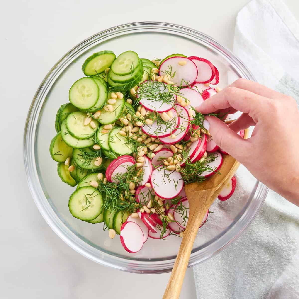 Woman adding fresh herbs to a bowl with cucumbers, radishes, and pine nuts.